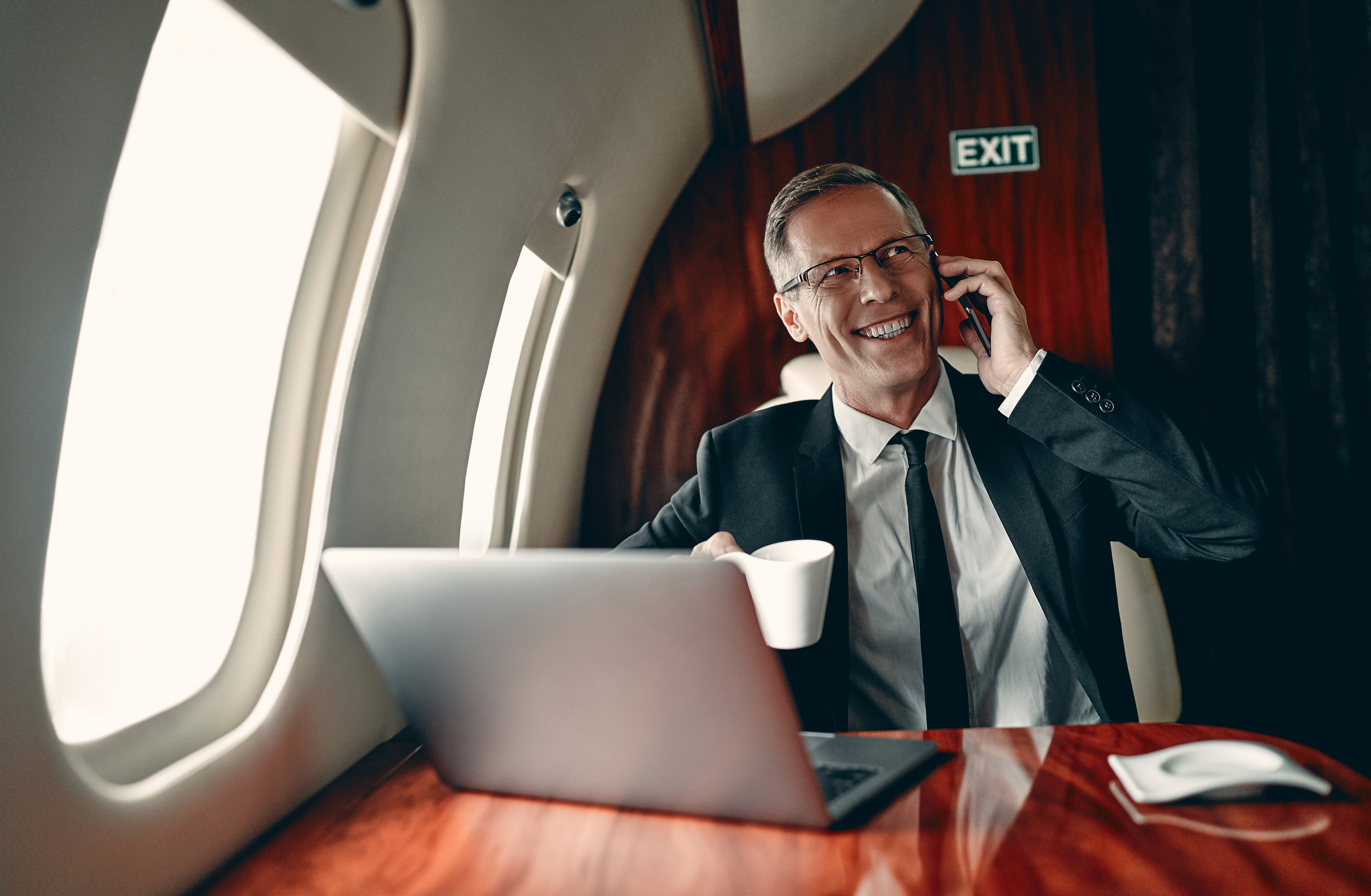 Senior businessman in suit is working on a laptop and speaking on mobile phone while flying in private jet.