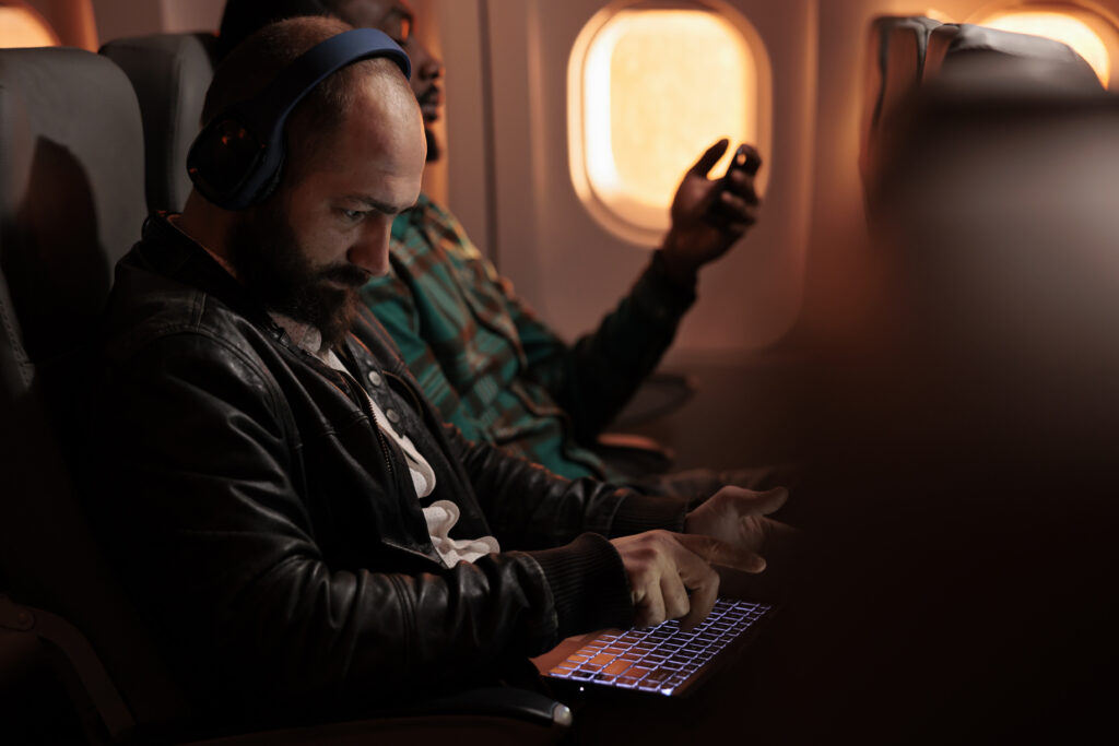Diverse group of passengers flying in economy class on plane jet, travelling to holiday destination. Using laptop and smartphone during sunset flight before arriving on vacation trip.